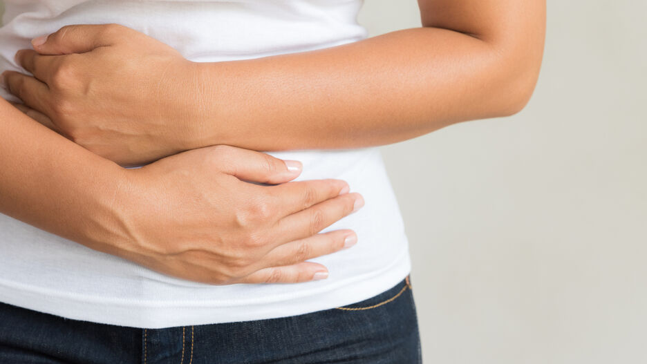 Abgebildet ist ein Torso einer Person in Jeans und weißem T-Shirt. Die Person hält sich mit den Armen den Bauch.