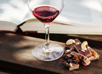 Ein Glas Wein steht vor einem geöffnetem Buch, daneben liegt Schokolade mit Nüssen