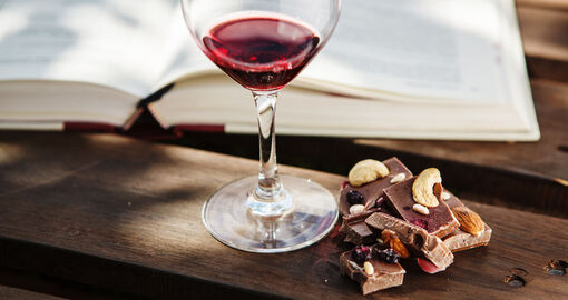 Ein Glas Wein steht vor einem geöffnetem Buch, daneben liegt Schokolade mit Nüssen