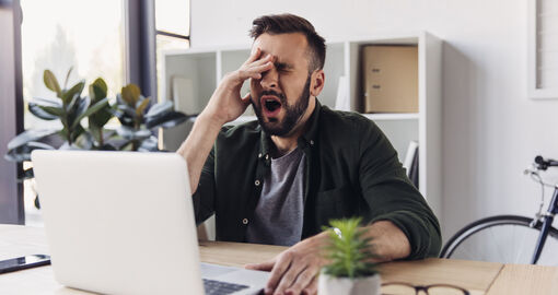 Ein Mann sitzt vor seinem Laptop am Arbeitsplatz und gähnt.