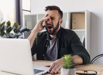 Ein Mann sitzt vor seinem Laptop am Arbeitsplatz und gähnt.