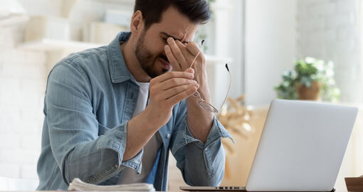 Mann im Jeanshemd sitzt augenzukneifend und Fingern an den Augen samt Brille in der anderen Hand vor dem Laptop.