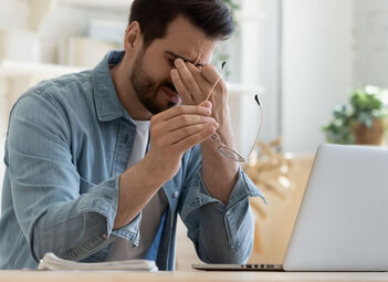 Mann im Jeanshemd sitzt augenzukneifend und Fingern an den Augen samt Brille in der anderen Hand vor dem Laptop.