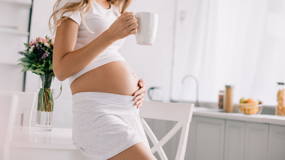 Schwangere Frau steht mit einer Tasse in der Küche-Ernährung in der Schwangerschaft - Schwangere dürfen Kaffee trinken. Sie sollten nur die Menge im Blick behalten.
